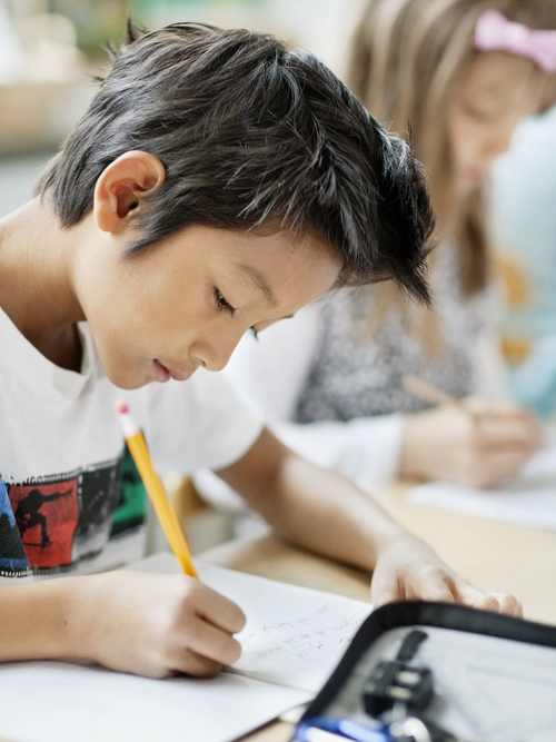 Bild på en elev bland sina klasskamrater, sittandes vid skolbänken med penna, papper och pennfodral framför sig.