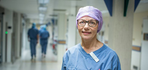 En person i blå operationskläder och lila hätta står i en sjukhuskorridor.