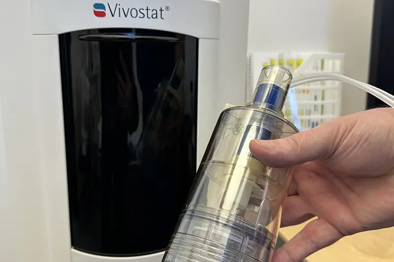 Närbild på en hand som håller i en plastbehållare intill en medicinteknisk apparat.
