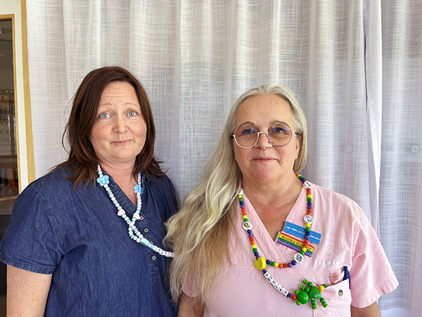 Två kvinnor i vårdpersonalkläder som står intill varandra och tittar in i kameran. 