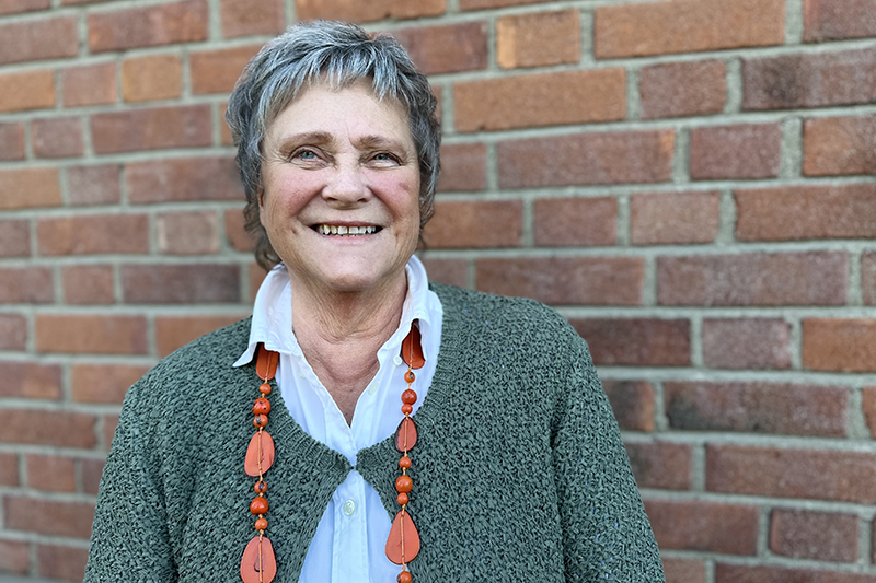 En person i grön kofta står vid en tegelvägg och ler mot kameran