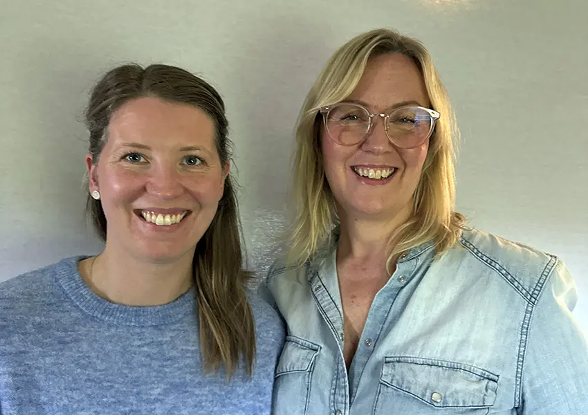 Två leende personer, en brunhårig med grå stickad tröja och en blond med glasögon och jeansskjorta