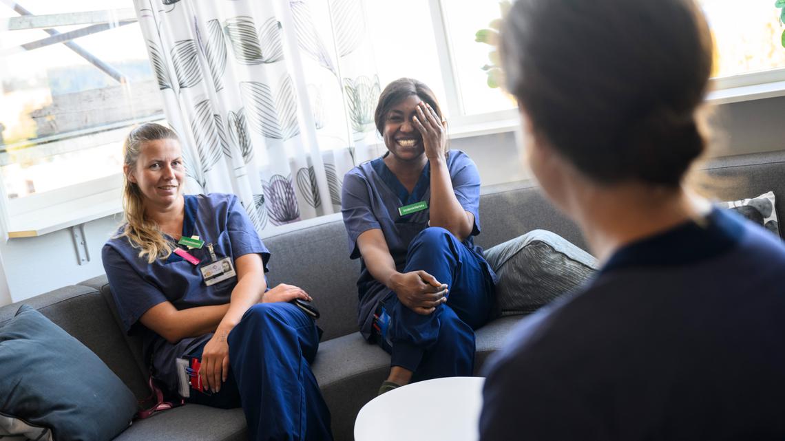 Två vårdklädda kollegor skrattar och ler i en soffa i ett personalrum, pratandes med en tredje kollega som skymtas i förgrunden.