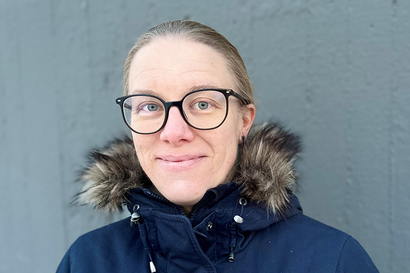 En person i mörkblå vinterjacka med pälskantad huva iförd glasögon och med ljust hår uppsatt i en tofs baktill tittar in i kameran