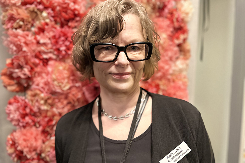 Person i bruna kläder och kort hår med glasögon. I bakgrunden en rosa rosentavla