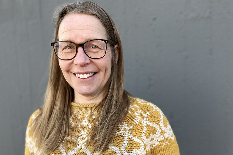 Bild på leende person med långt brunt hår och glasögon iklädd en stickad tröja i gult och vitt.