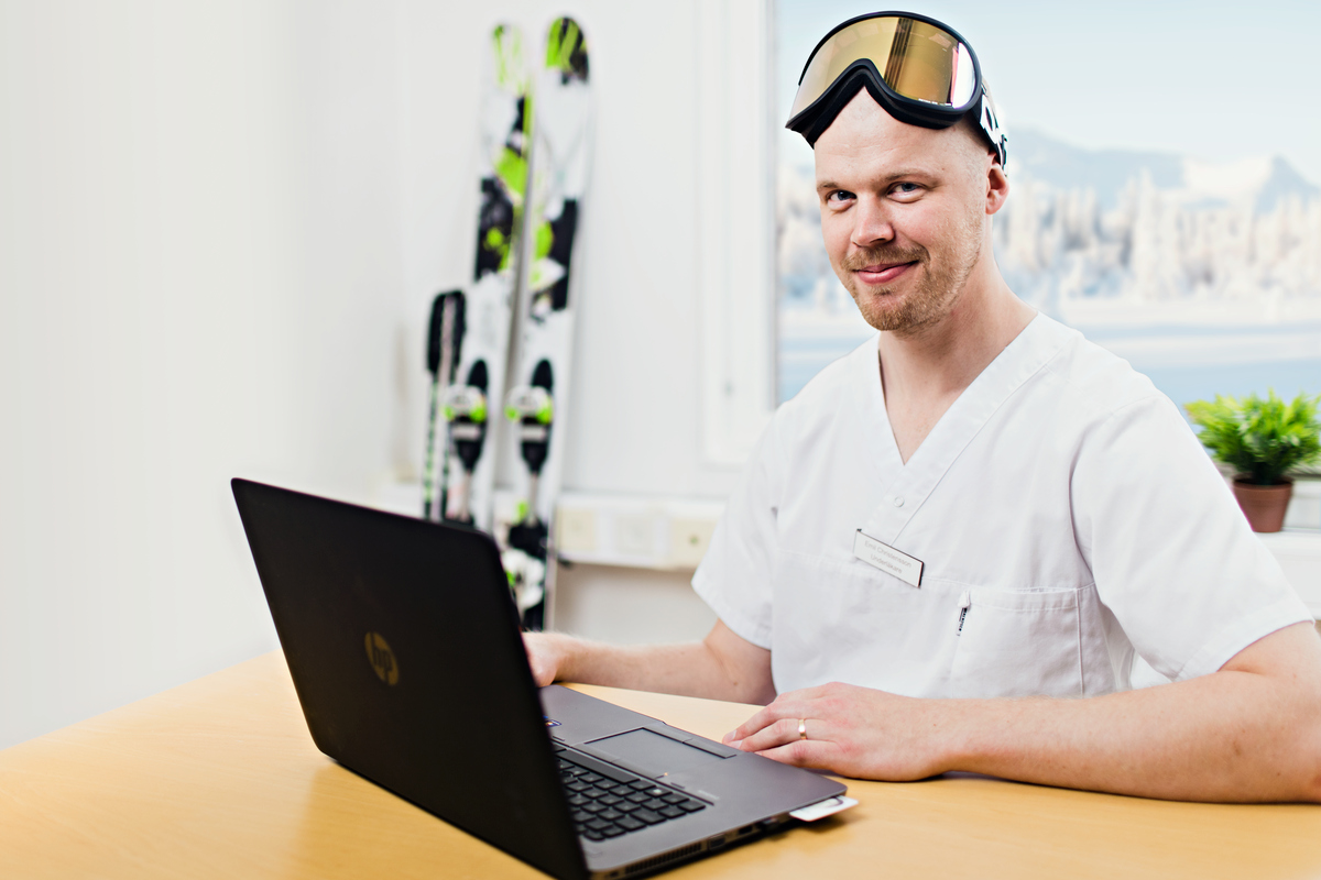 Läkare med slalomglasögon på sig och samtidigt jobbar vid en bärbar dator. I bakgrunden syns slalomskidor och i fönstret ett gnistrande snölandskap.