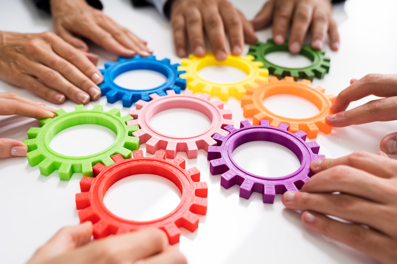 Flera händer runt en vit bordsskiva, håller i olika färgade plastcirklar för att illustrera kugghjul/växlar i ett samarbete.