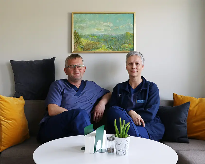 Två vårdklädda yrkespersoner sitter i en soffa i ett personalrum, med färgglada kuddar runt om och en tavla ovanför.