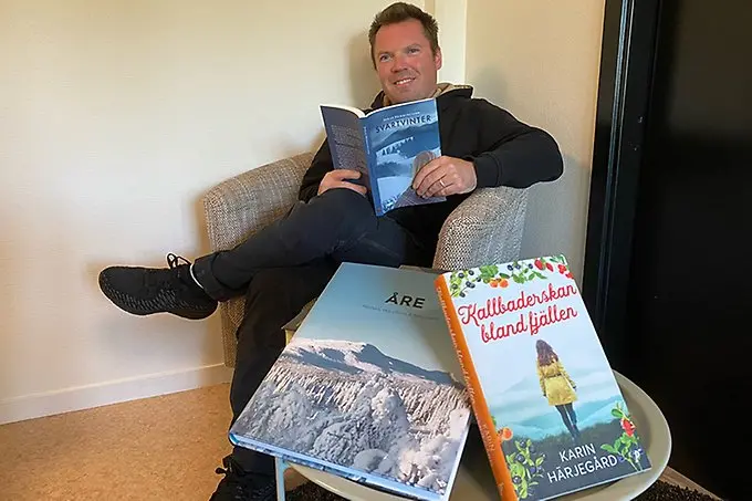 En person sitter i en fåtölj med en öppen bok i händerna. Personen tittar in i kameran och ler. 