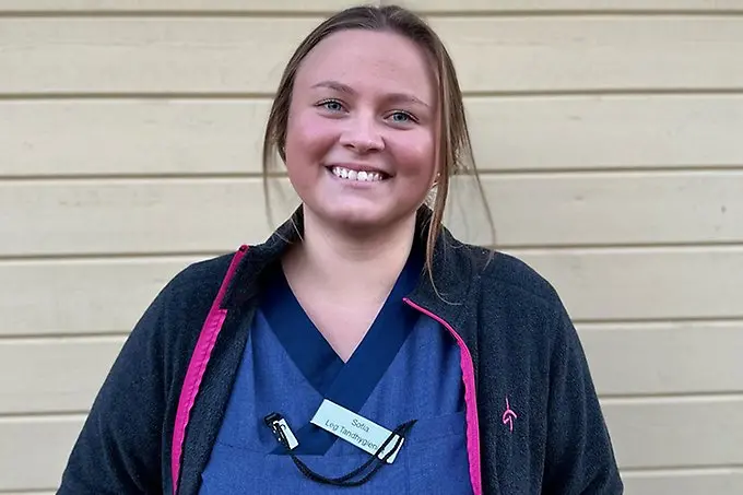 En person står mot en ljus trävägg och ler brett in i kameran.