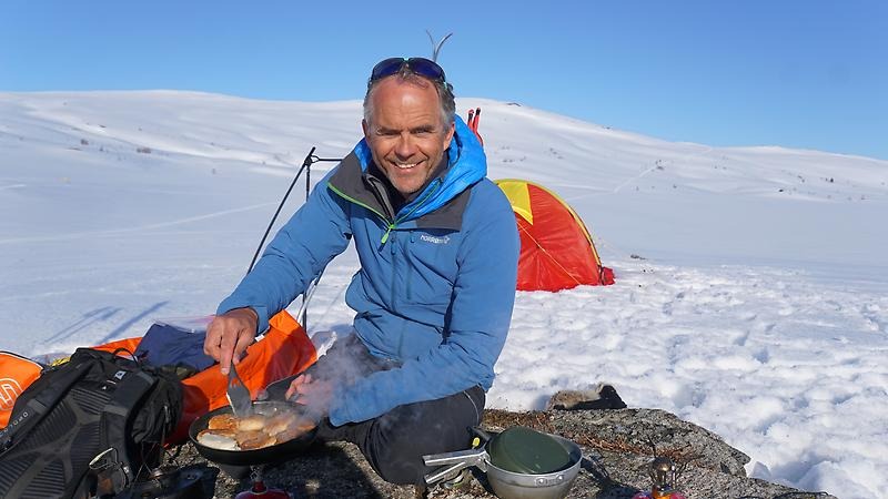 Företagaren Tore Holand sitter vid en snöig klippa och lagar mat på sitt stormkök. I bakgrunden syns snöklädda fjäll och ett vintertält.
