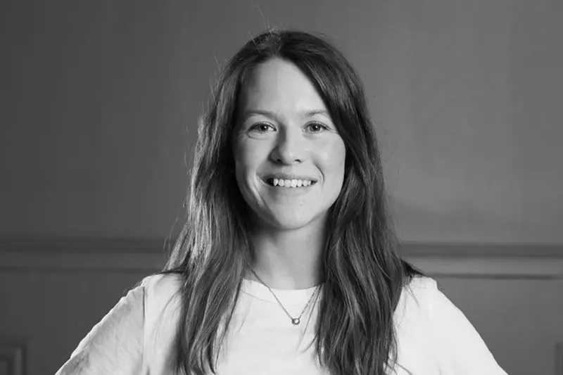 Svartvitt foto på en leende person med långt mörkt hår och vit långärmad tröja.