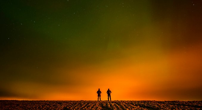 Siluetterna av två små personer som står och tittar mot en delvis orange och delvis mörk himmel.