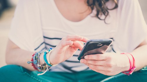 Kvinna sitter i skräddarställning och tittar i sin mobiltelefon. Man ser inte ansiktet utan bara händerna och kroppen.