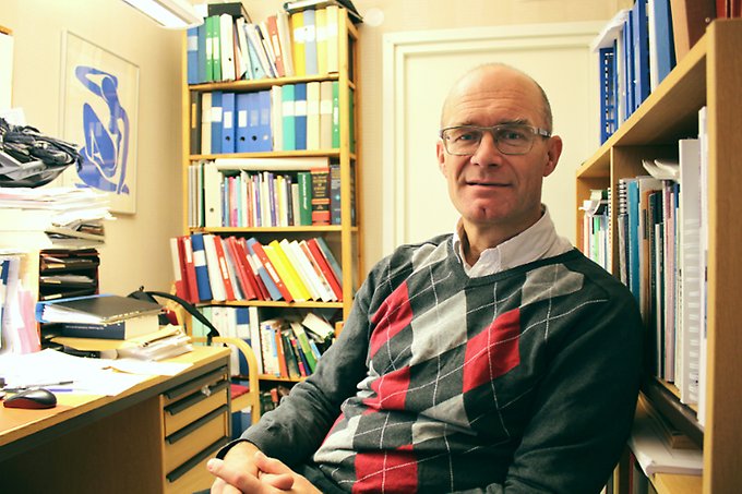 en man sitter tillbakalutad på en kontorsstol framför ett skrivbord och bokhyllor med pärmar och böcker