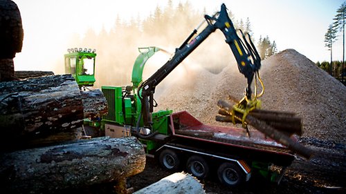En skogsmaskin lägger upp timmer från stora timmerhögar på ett timmerflak.