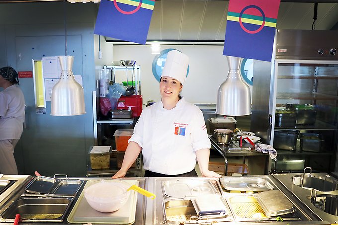 en kvinnlig kock står bakom en matdisk med kantiner fyllda av mat, samiska vimplar hänger ovanför henne