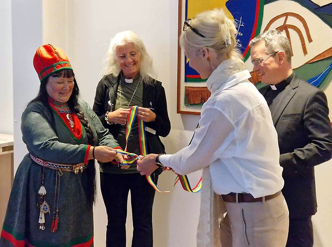 Fyra personer står framför ett samiskt konstverk varav den ena, en kvinna i samisk högtidsdräkt knyter samman ett tygband i de samiska färgerna.