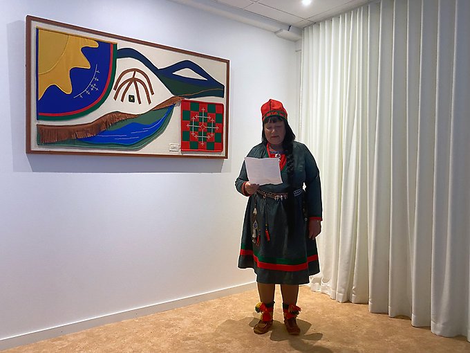 En kvinna i samiska högtidskläder läser ur ett papper framför ett samiskt konstverk