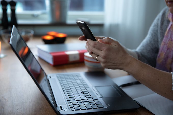 En person sitter vid ett bord och håller en mobiltelefon i händerna. På bordet står det en bärbar dator. 