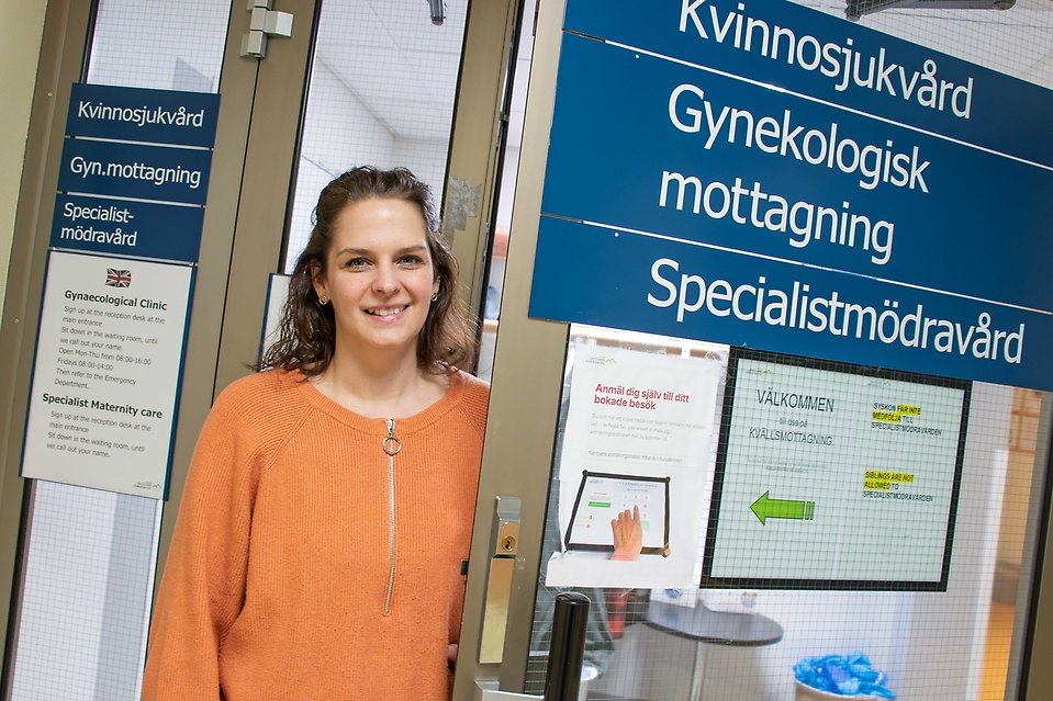 En person med axellångt brunt hår och klädd i orange tröja håller upp en dörr med skyltarna kvinnosjukvård, gyneokologisk mottagning och specilistmödravård.