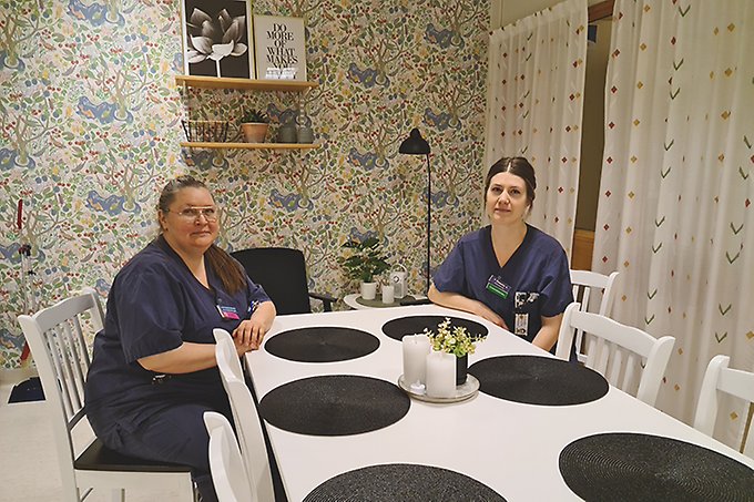 Två personer i blå vårdkläder sitter mittemot varandra i ett kök vid ett vitt matbord