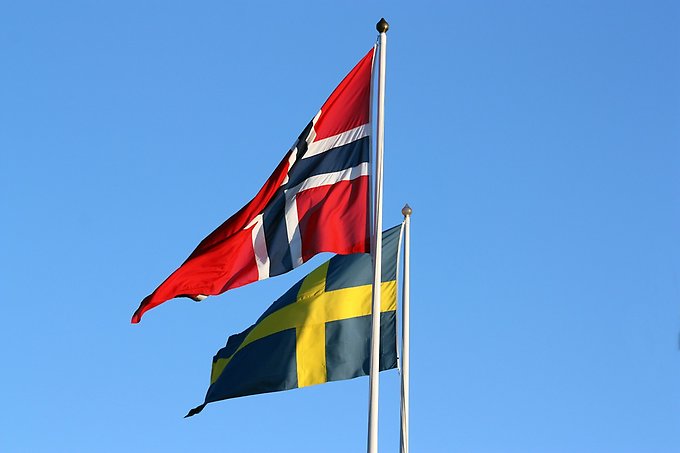 Närbild på en norsk och en svensk flagga som vajar i varsin flaggstång mot bakgrund av en blå himmel.