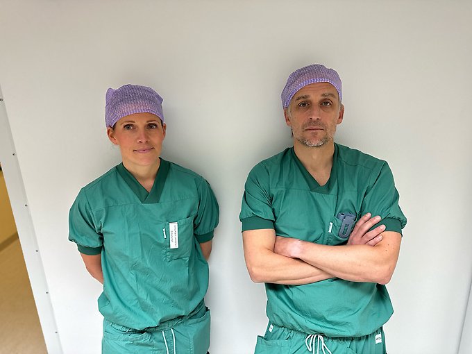 Två personer iklädda gröna operationskläder och mössor står mot en vit vägg och tittar in i kameran
